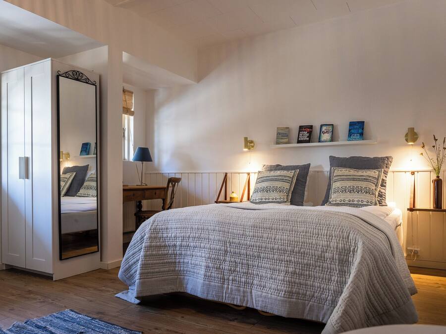 A bedroom with a large bed, a bedside table, a chair, a desk, a mirror, and a shelf with books.