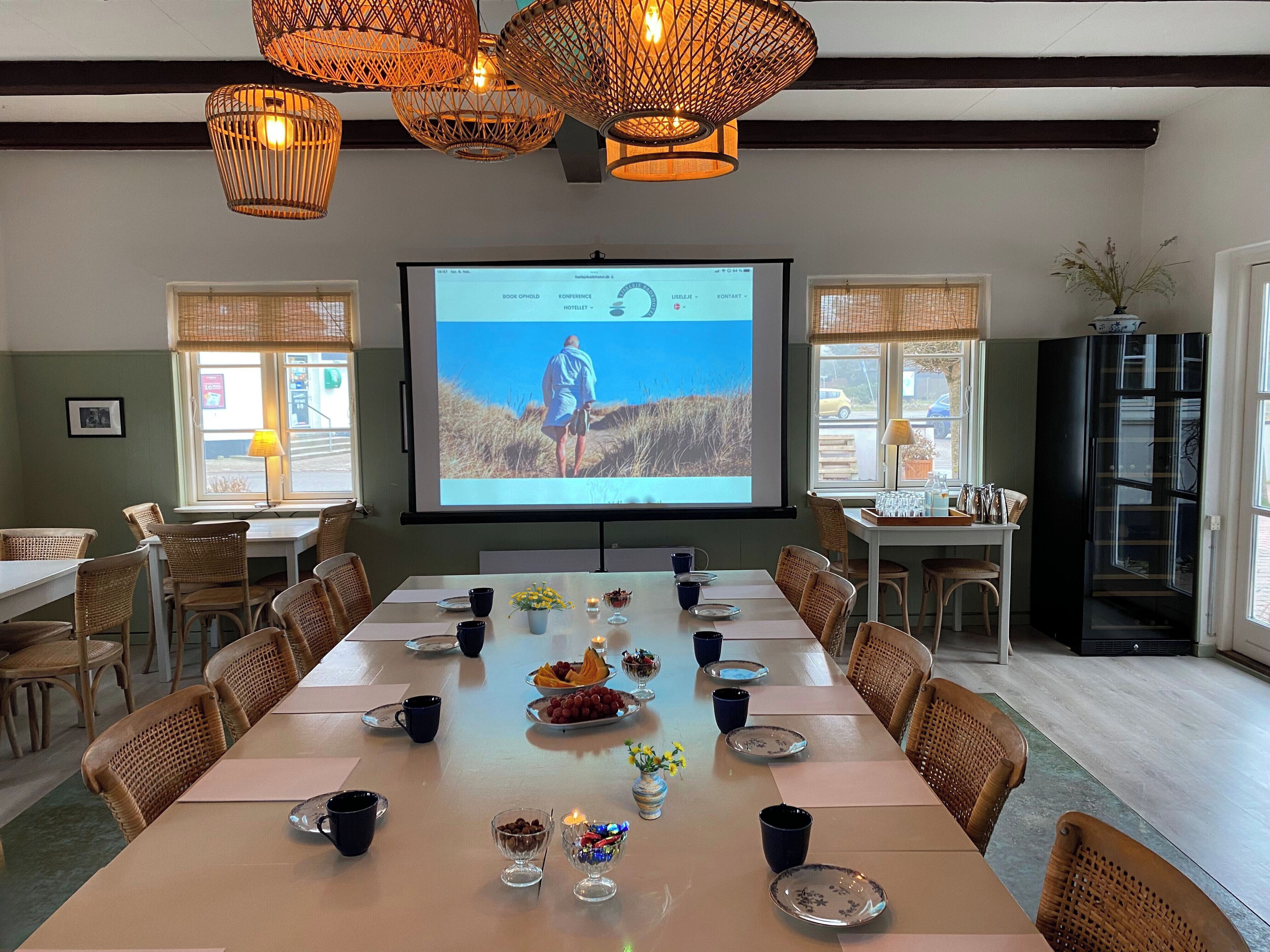 A conference room with a large screen displaying a presentation, wicker chairs, a long table set for a meeting, and a view of the outdoors through the windows.