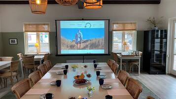 A conference room with a large screen displaying a presentation, wicker chairs, a long table set for a meeting, and a view of the outdoors through the windows.