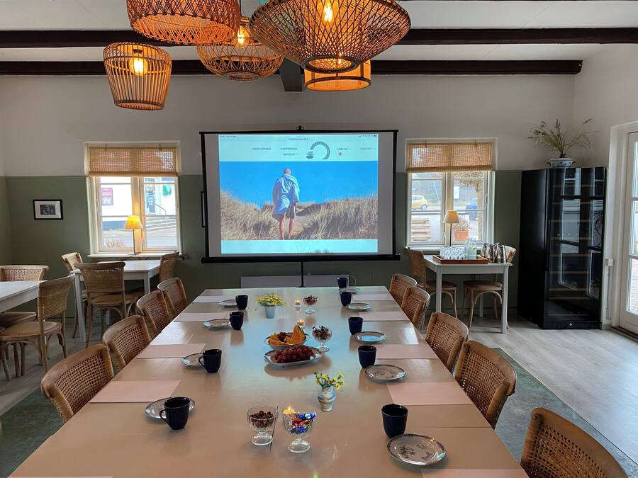 Un salón de conferencias con una pantalla grande mostrando una presentación, sillas de mimbre, una mesa larga dispuesta para una reunión y vistas al exterior a través de las ventanas.