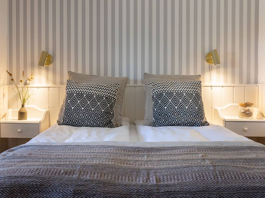 Una cama bien hecha con almohadones estampados, una mesita de noche con un florero y una pared con rayas verticales.