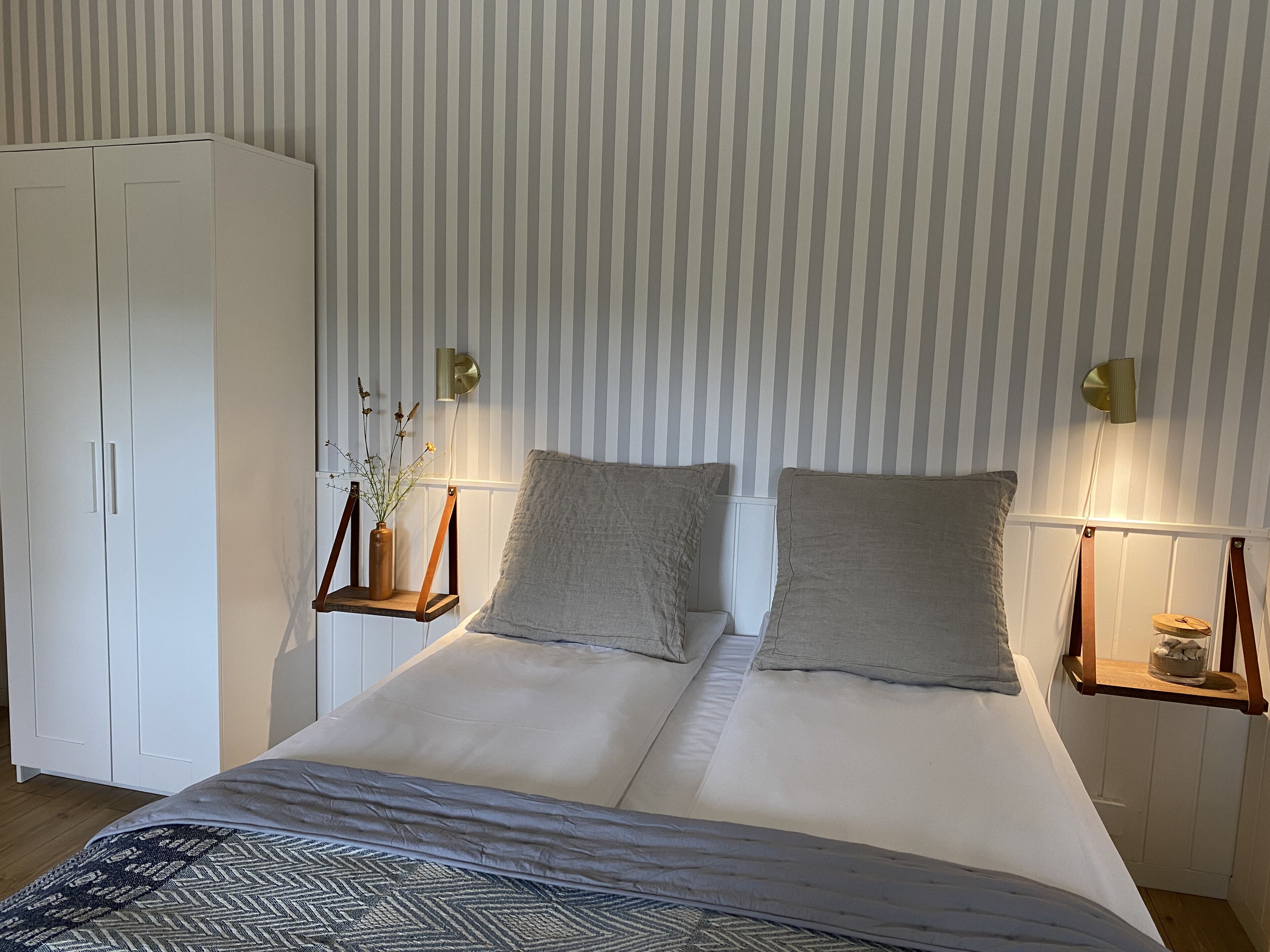 A double bed with a patterned throw, two bedside tables with lamps, a wardrobe, and a striped wall.