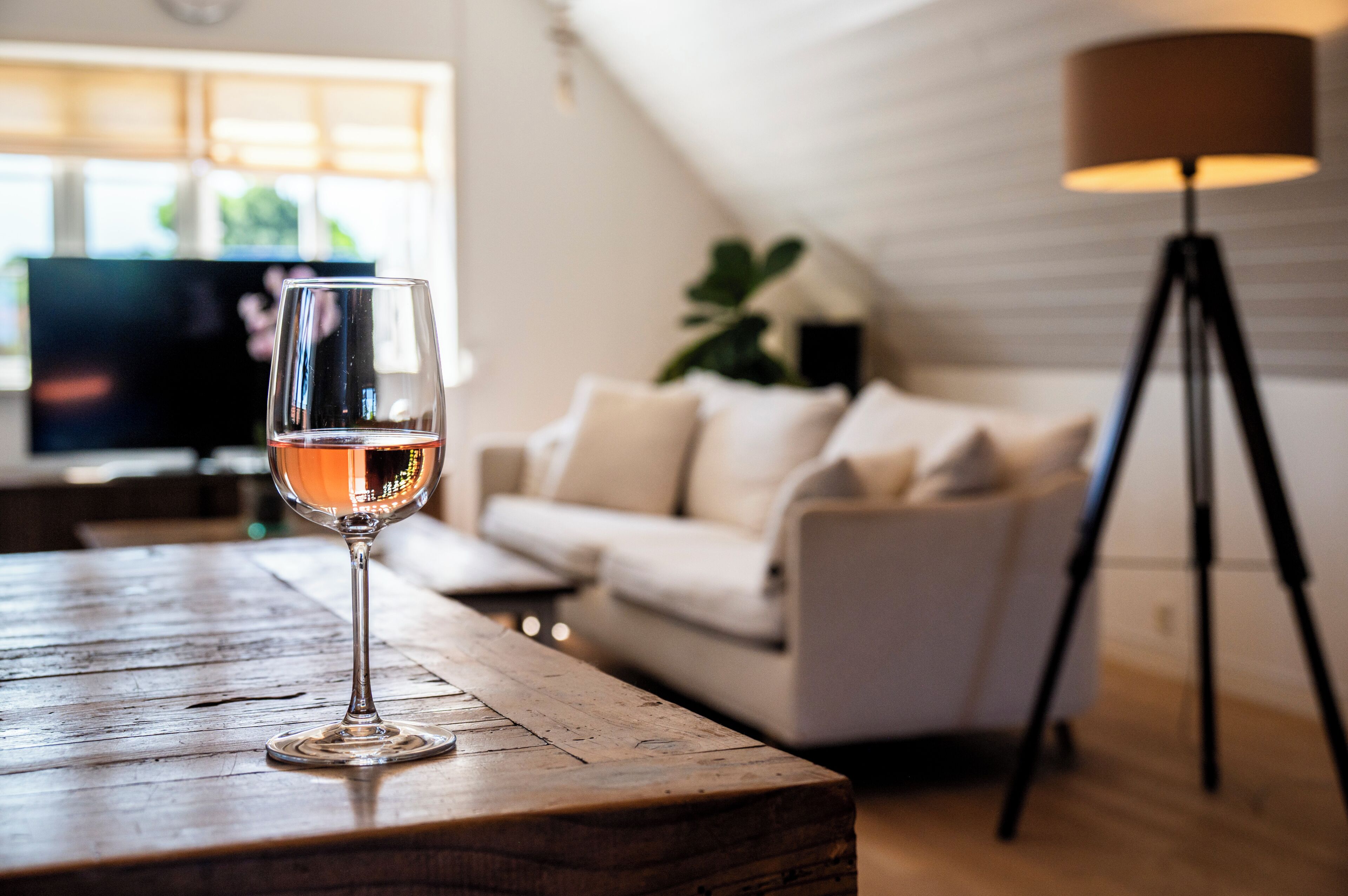 Una copa de vino sobre una mesa de madera en una habitación con un sofá, una lámpara y un televisor al fondo.