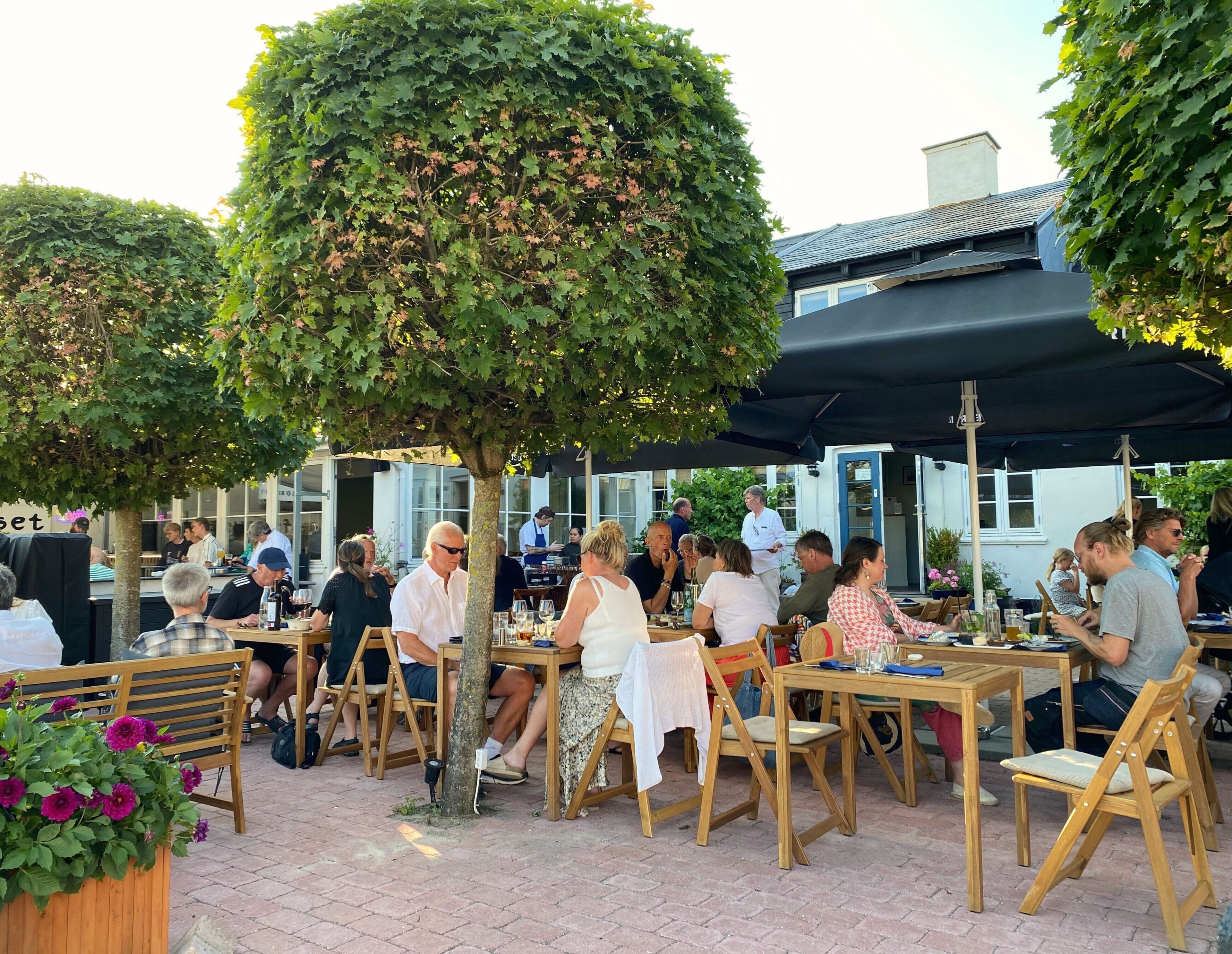 An outdoor dining area with wooden tables and chairs, surrounded by trees and covered by a black canopy.