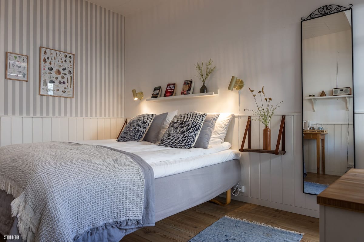 Quarto com cama, mesa de cabeceira, espelho e prateleiras na parede com objetos decorativos.