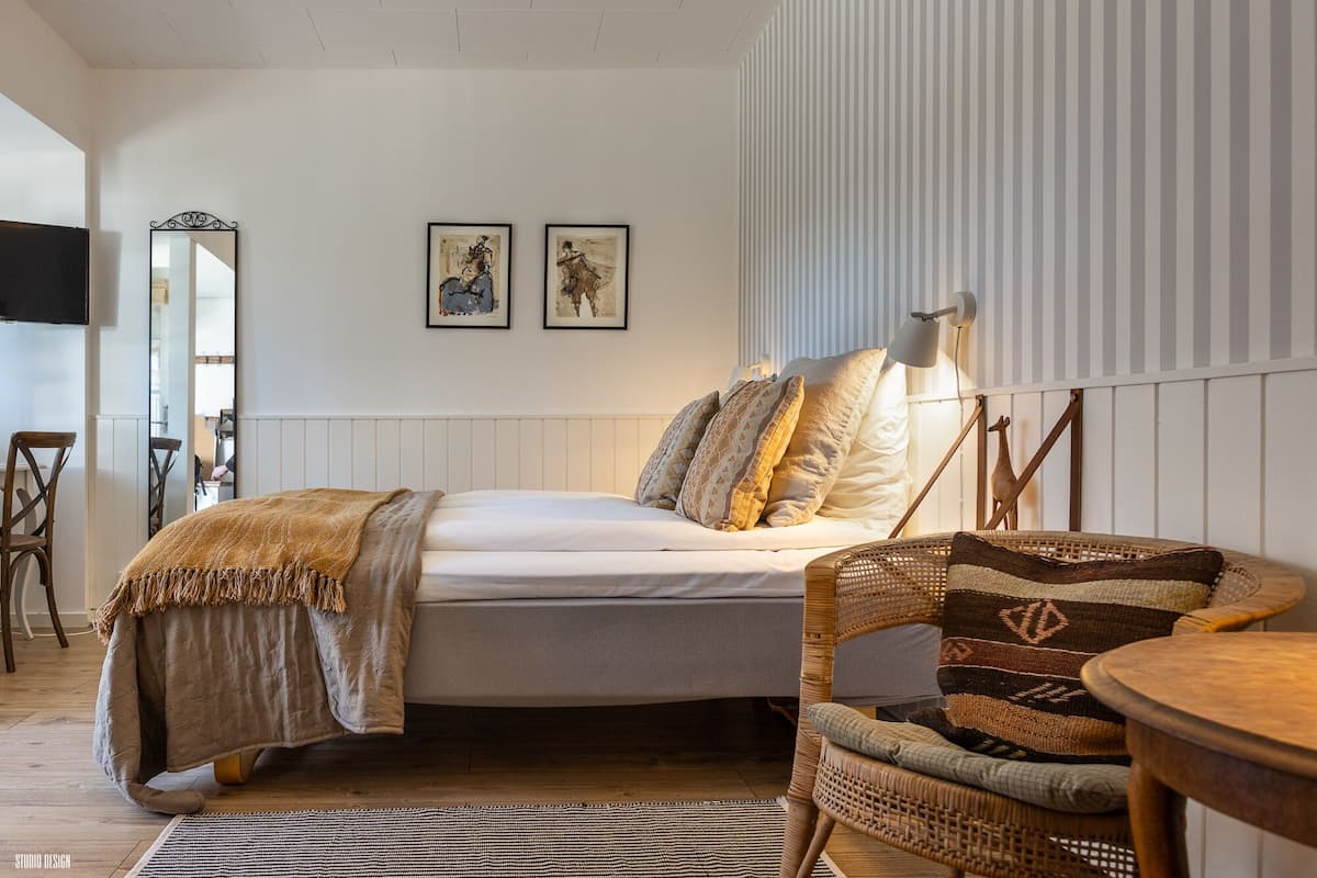 Um quarto aconchegante com cama, poltrona, mesa e espelho. Há duas imagens emolduradas na parede.