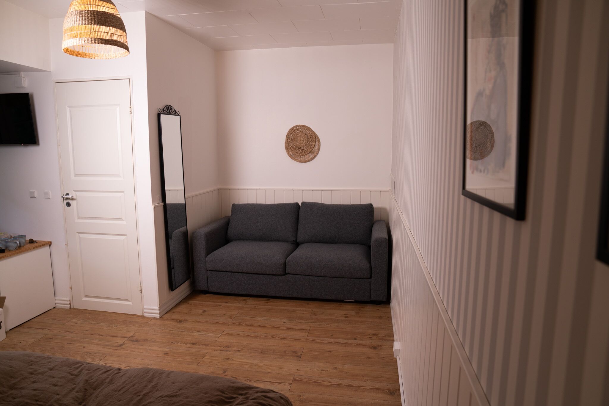 Ein Zimmer mit grauem Sofa, weißer Tür, Flachbildfernseher und einer gewebten Leuchte.