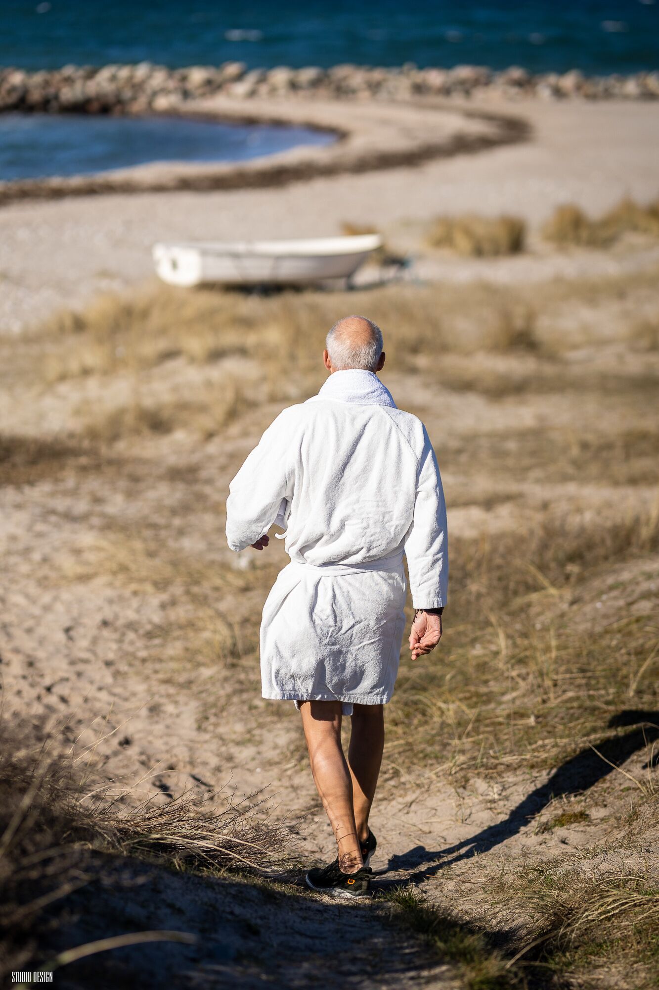 A person in a white robe walking on a sandy path with a body of water and a boat in the background.