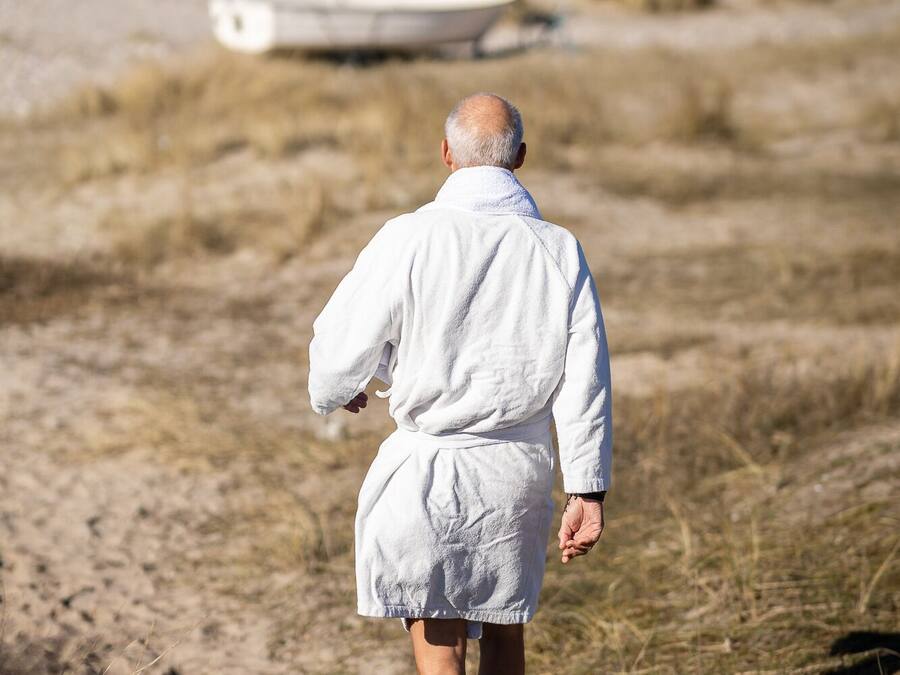 Une personne vêtue d’une robe blanche marchant sur un sentier sablonneux, avec une étendue d’eau et un bateau en arrière-plan.