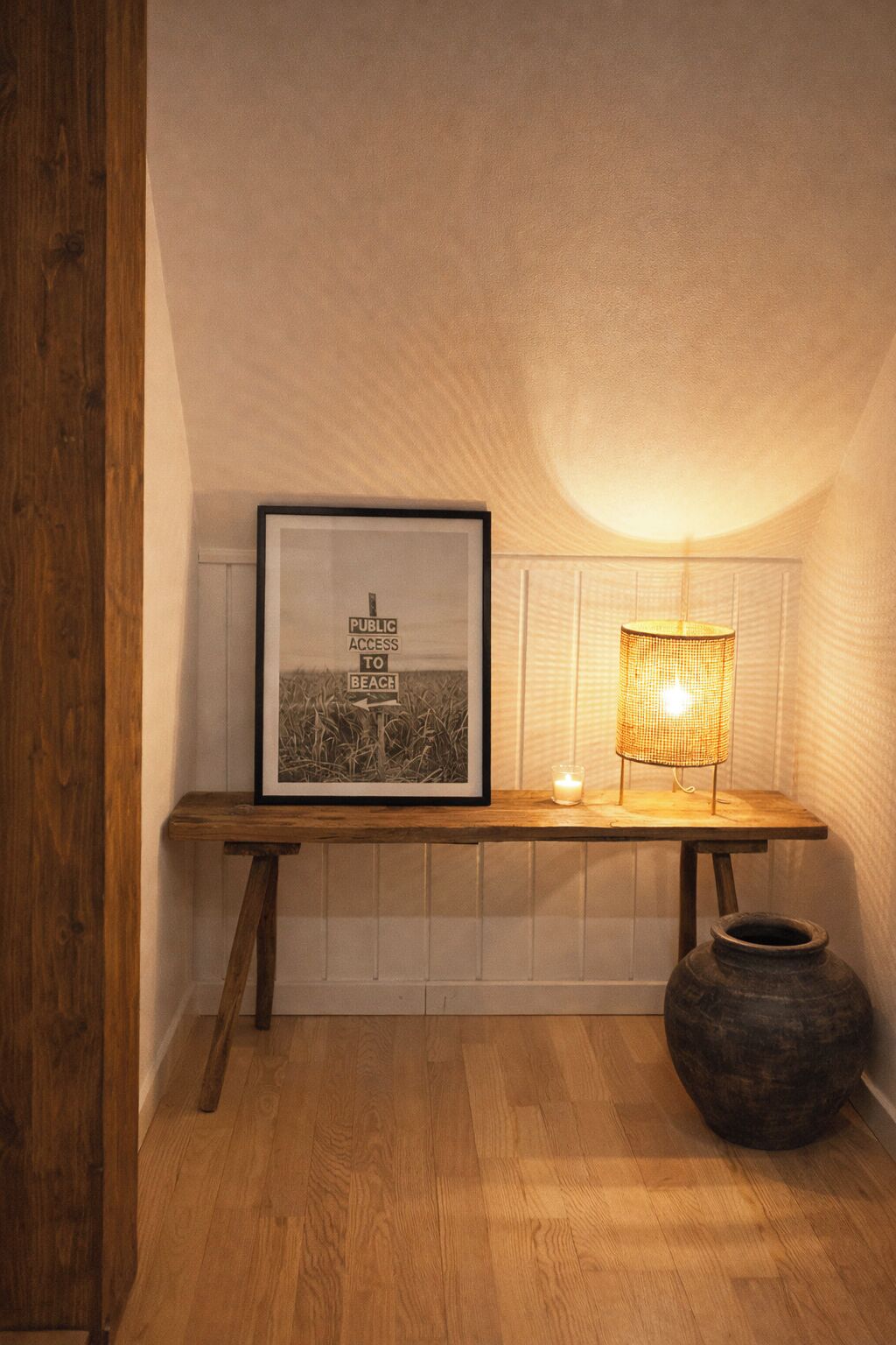 A framed sign with 'PUBLIC ACCESS TO BEACH' text, a table with a candle and a lamp, and a large pot on the floor.