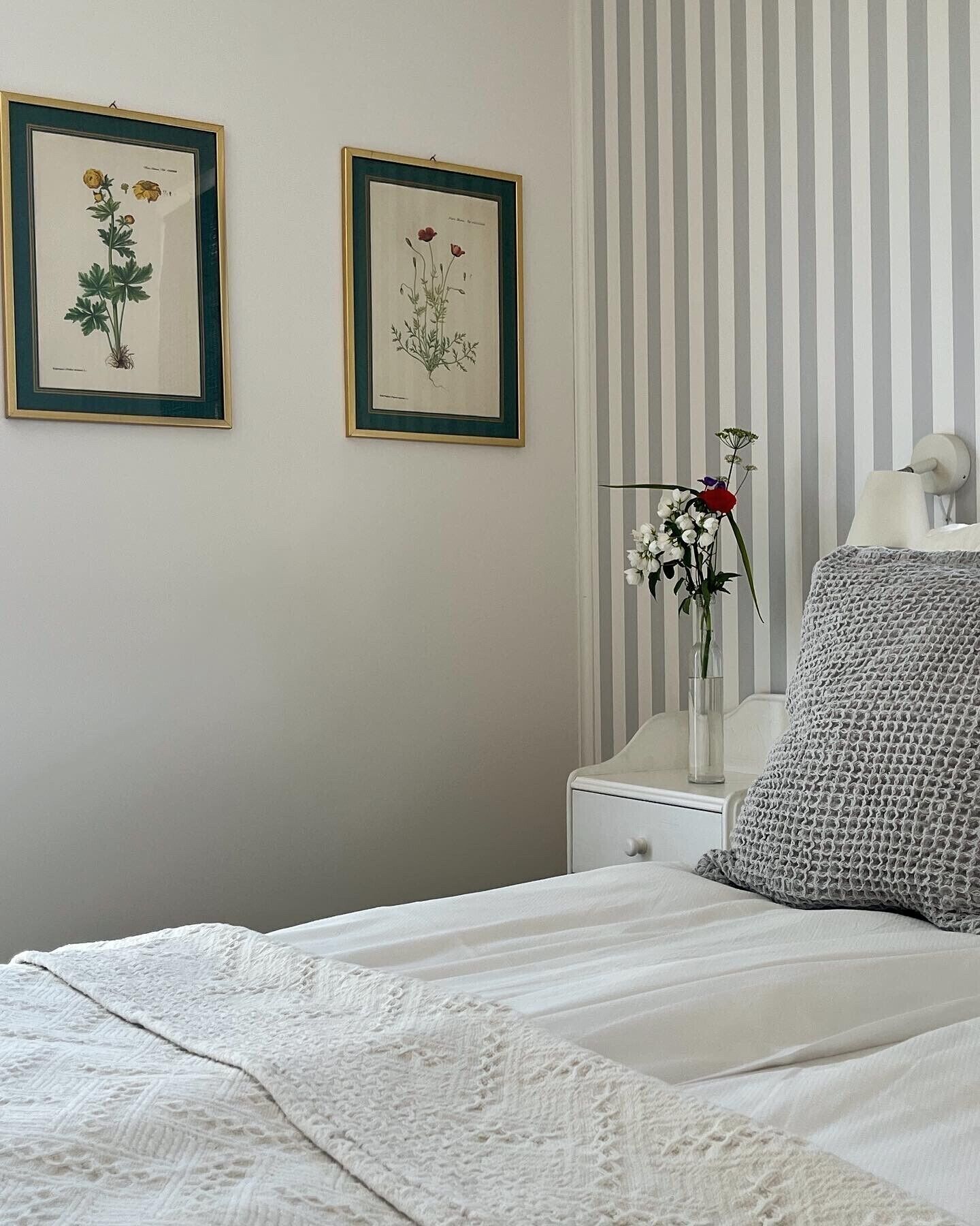 Una camera da letto con un letto, un comodino, una lampada e due stampe incorniciate con motivi floreali alle pareti.