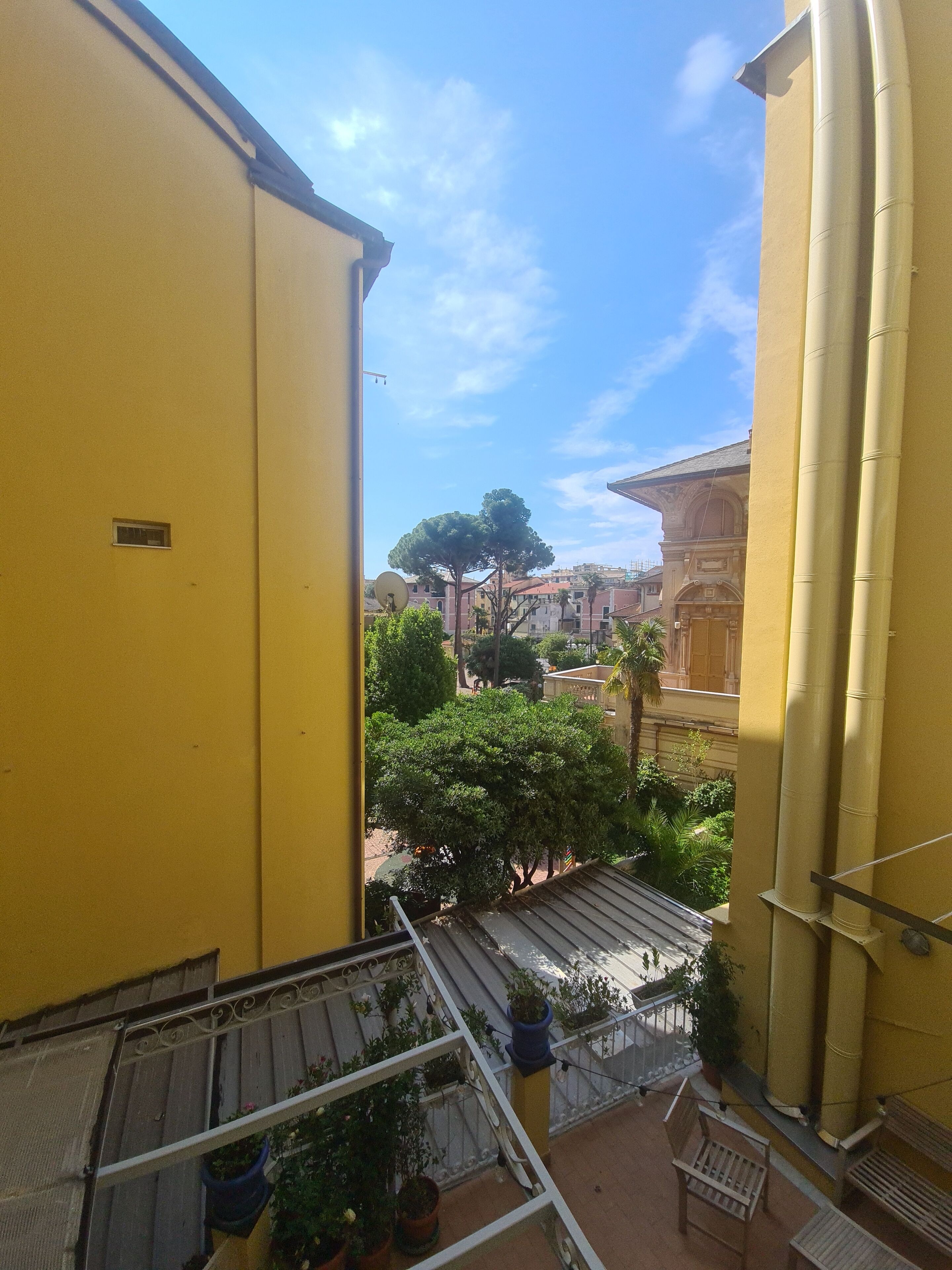 A yellow building with a balcony overlooking a courtyard with trees and a distant building.