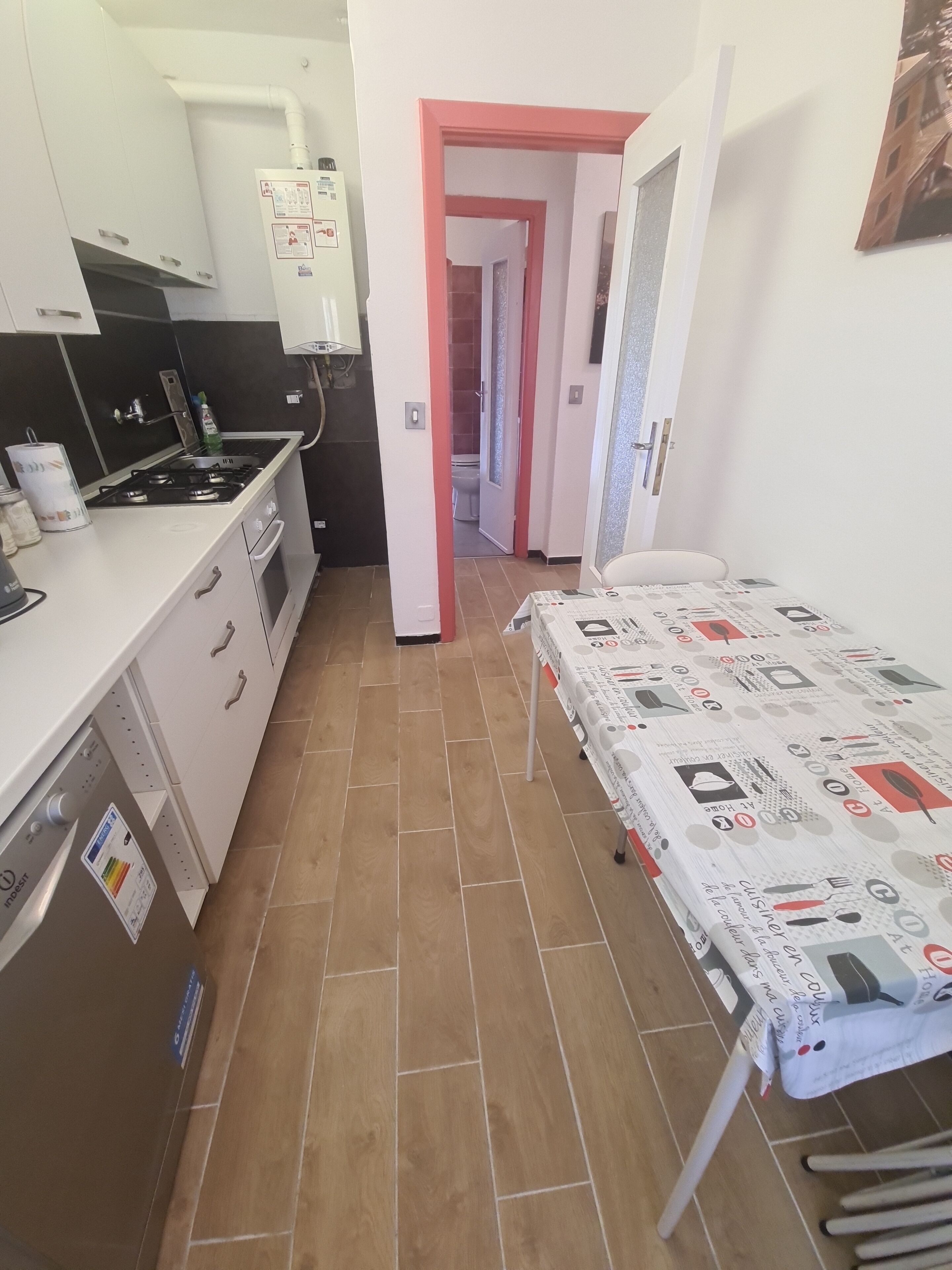 A compact kitchen with a gas stove, white cabinets, and a tiled floor. A dining table with a patterned tablecloth is visible in the foreground.
