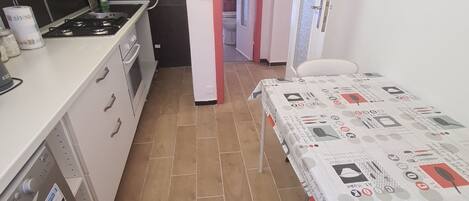 A compact kitchen with a gas stove, white cabinets, and a tiled floor. A dining table with a patterned tablecloth is visible in the foreground.
