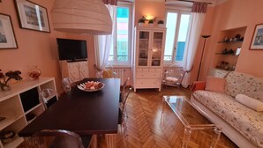 Ein gemütliches Wohnzimmer mit Holzdielen im Fischgrätmuster, einem Esstisch mit Stühlen, einer Couch, einem Fernseher und einem Schrank mit Glastüren.
