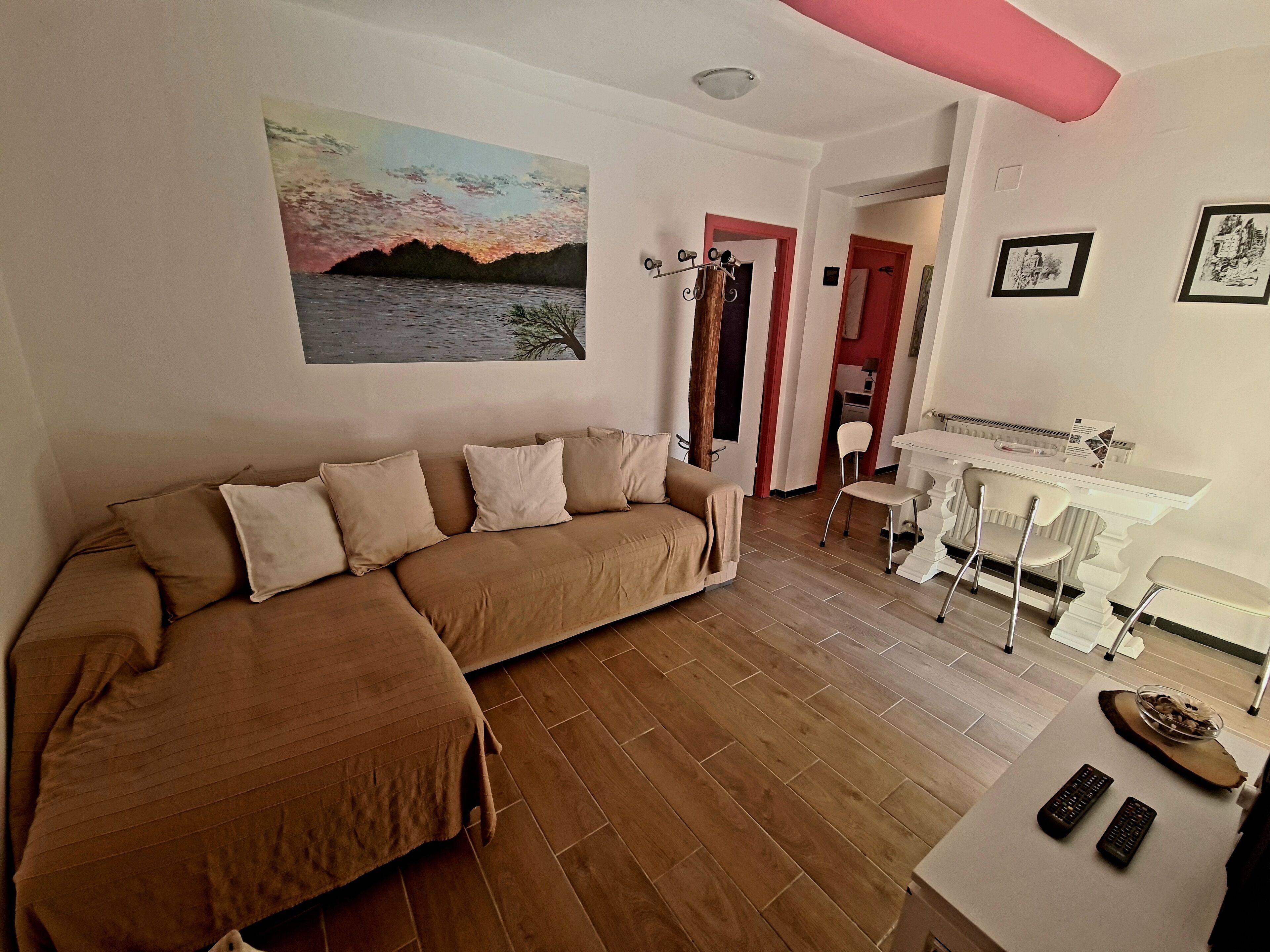 A living room with a brown sofa, a painting of a sunset, a dining area with a white table and chairs, and a kitchen in the background.