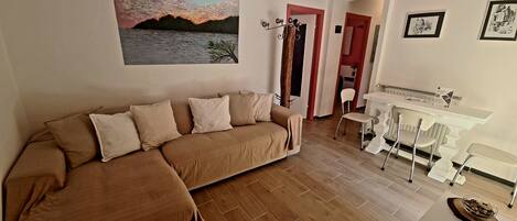 A living room with a brown sofa, a painting of a sunset, a dining area with a white table and chairs, and a kitchen in the background.