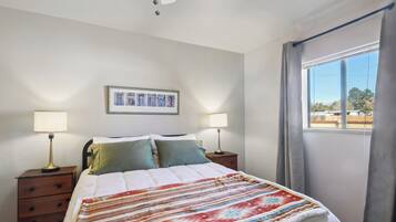 Un dormitorio con cama, dos mesitas de noche con lámparas, un cuadro en la pared y una ventana con cortinas.