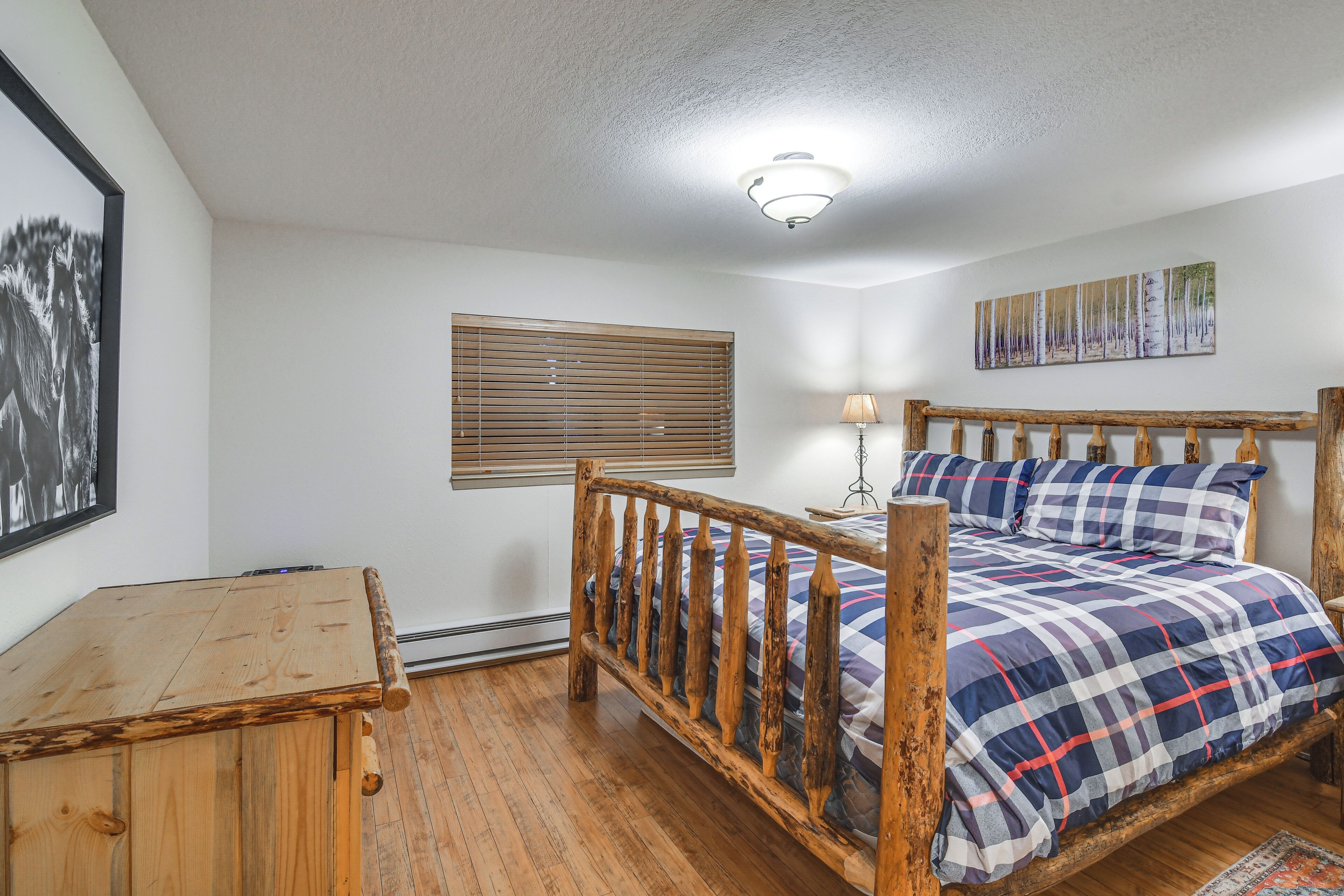 Un dormitorio con estructura de cama de madera, mesita de noche de madera, ventana con persianas, lámpara de pared y un cuadro enmarcado.