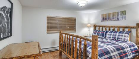 Un dormitorio con estructura de cama de madera, mesita de noche de madera, ventana con persianas, lámpara de pared y un cuadro enmarcado.