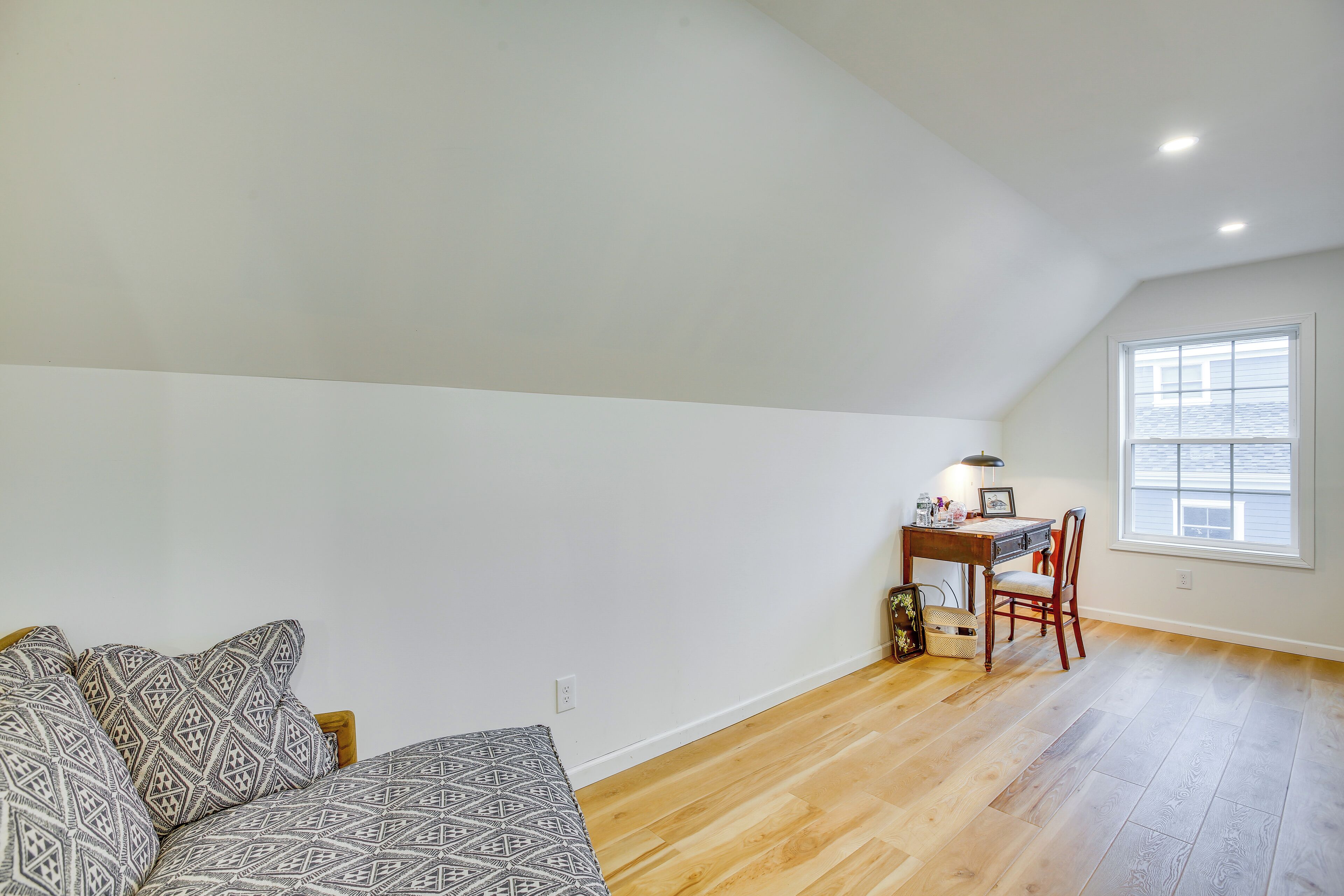 A room with a wooden floor, a white wall, a desk with a chair, and a patterned sofa.