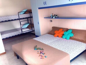 Un dormitorio con literas, una cama con ropa de cama estampada, una mesita con objetos dispersos y un estante con adornos.