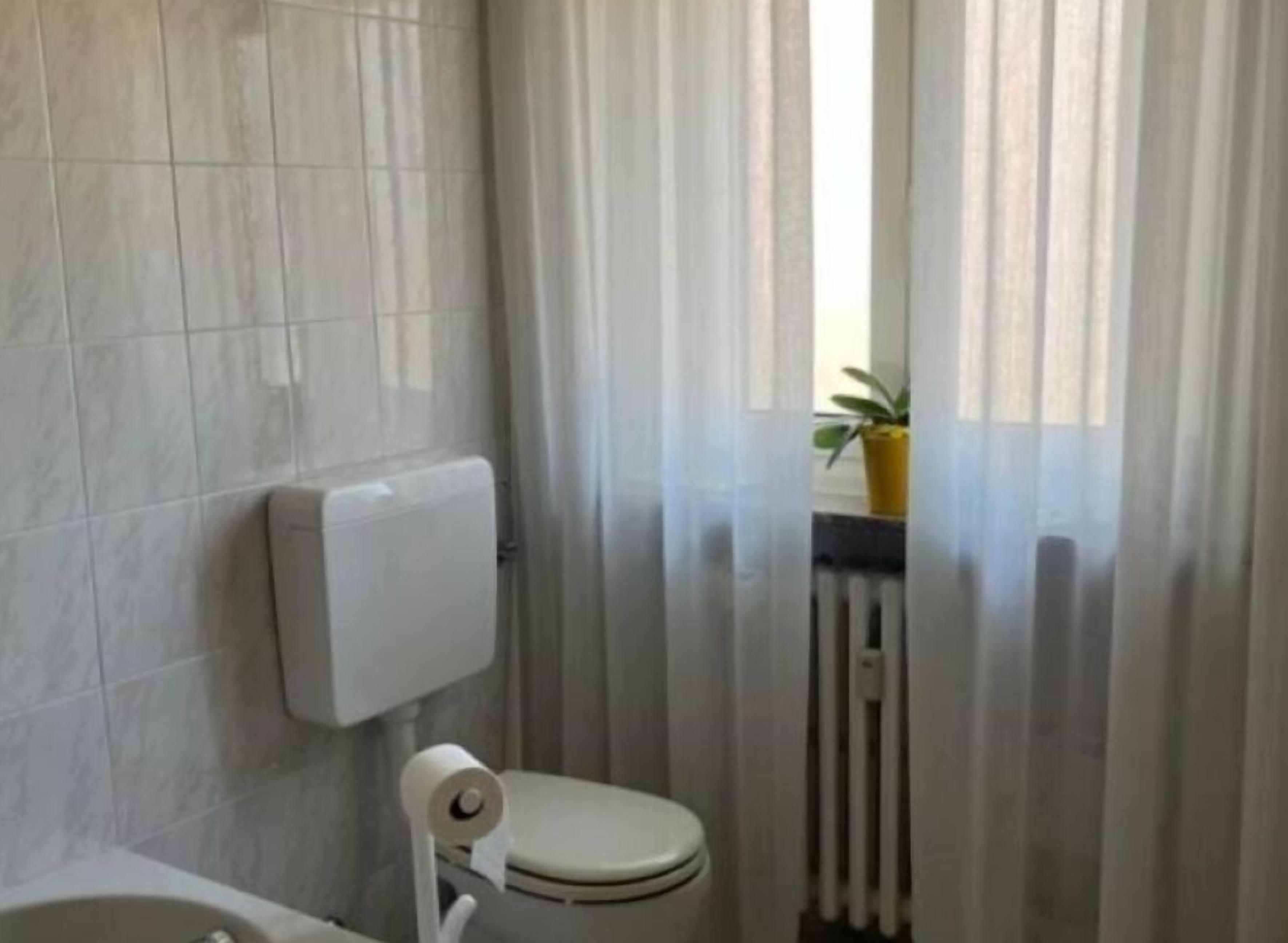 A bathroom with a toilet, a roll of toilet paper, a white toilet paper holder, a radiator, and a window with curtains.