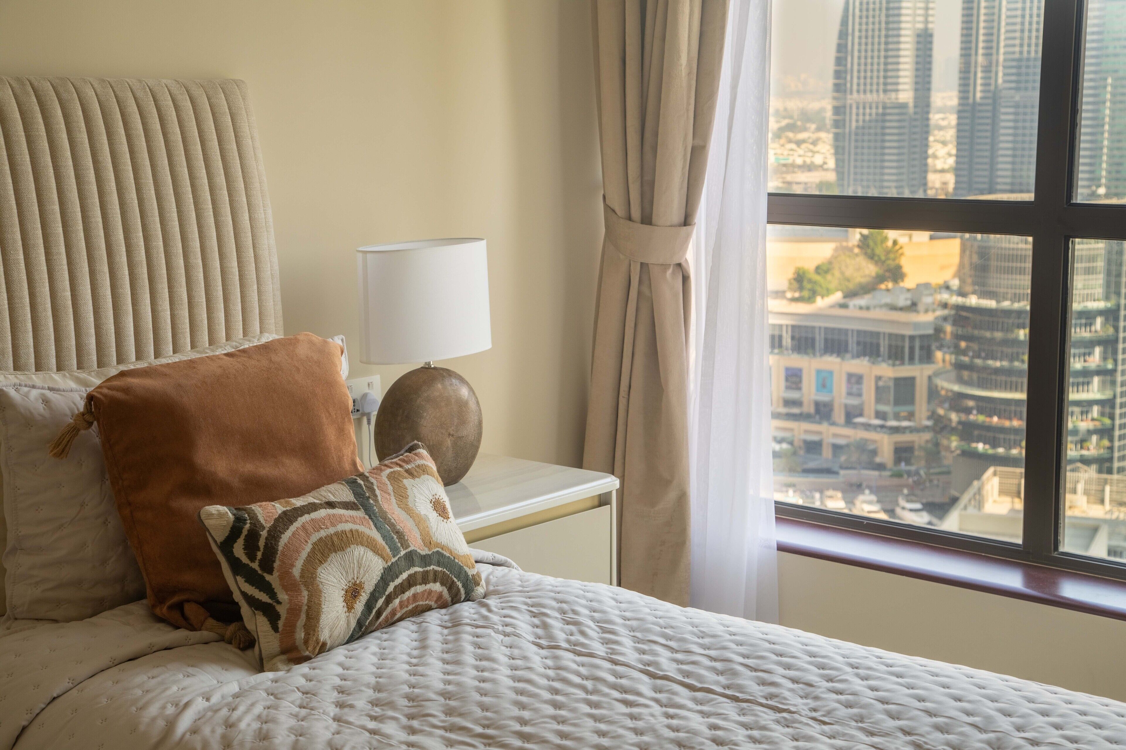 A hotel room with a bed, a nightstand, a lamp, and a view of a cityscape through the window.