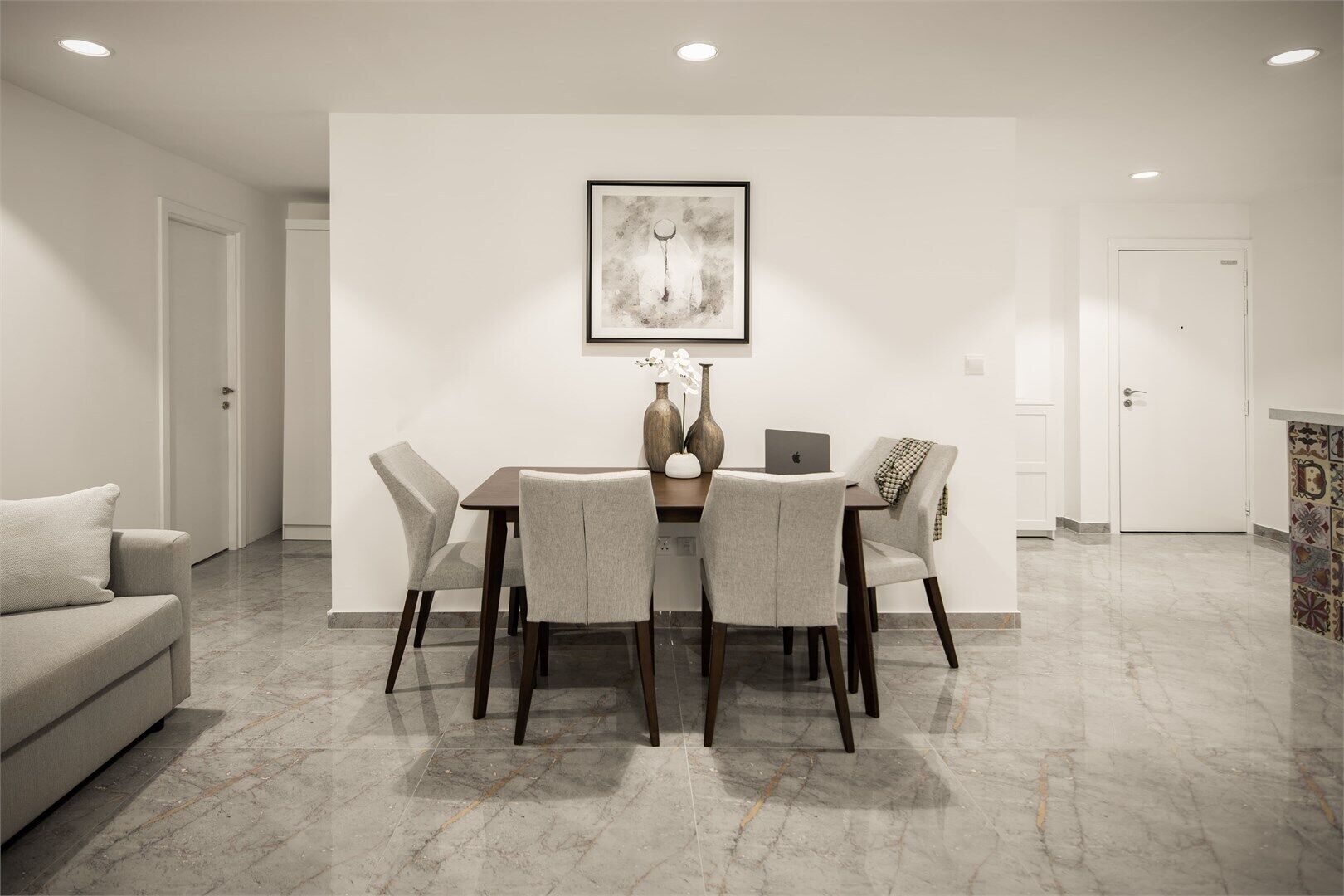 Un comedor moderno con piso de mármol, una pared blanca con un cuadro abstracto enmarcado y un ordenador portátil sobre la mesa.
