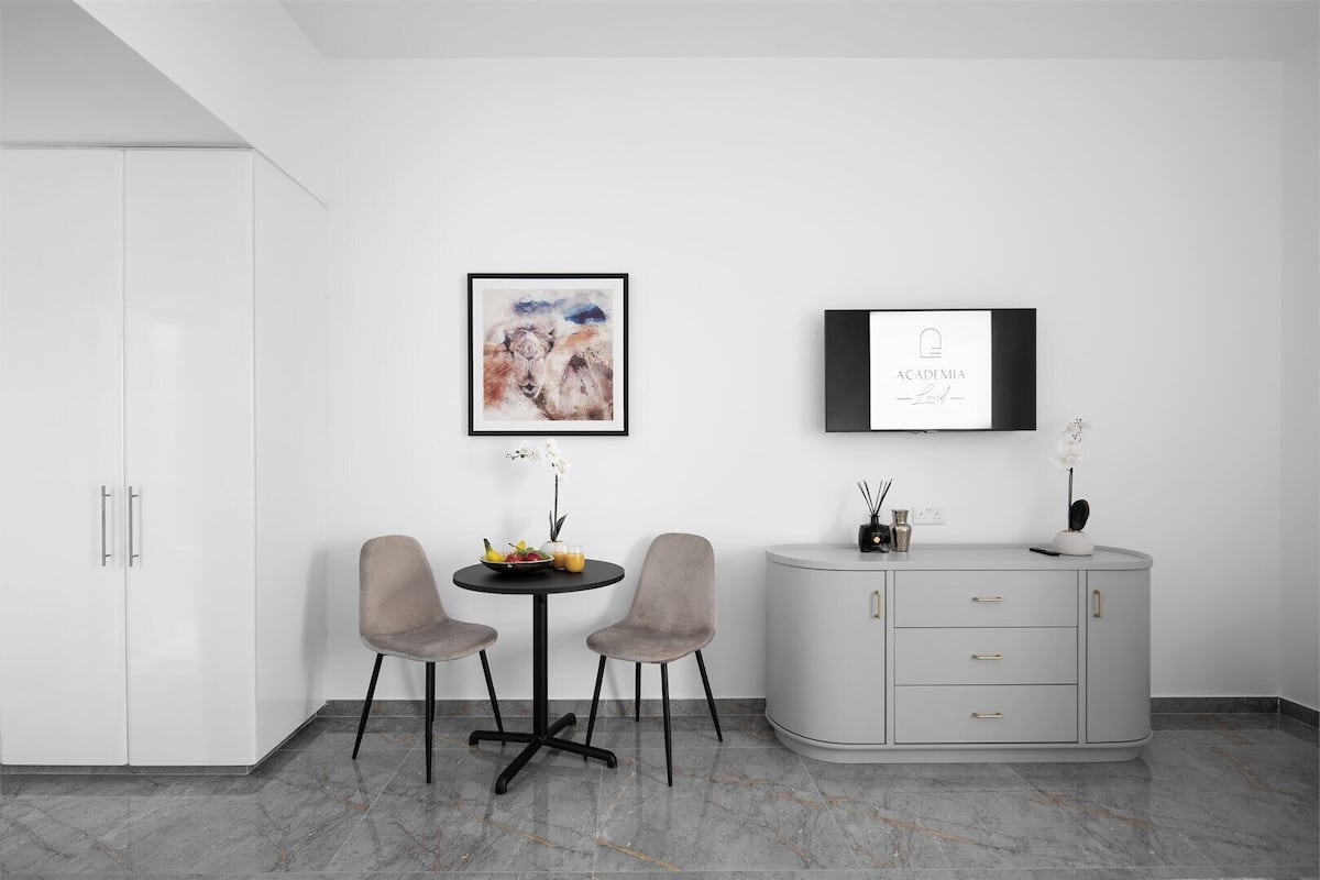 A modern room with a white wardrobe, a grey cabinet, and two grey chairs around a small round table with a fruit bowl and a vase of flowers.