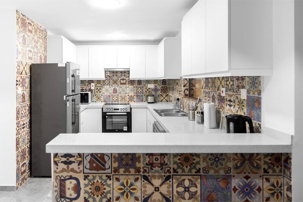 A modern kitchen with white cabinets, a central island, a microwave, an oven, and a sink. The backsplash features colorful, patterned tiles.