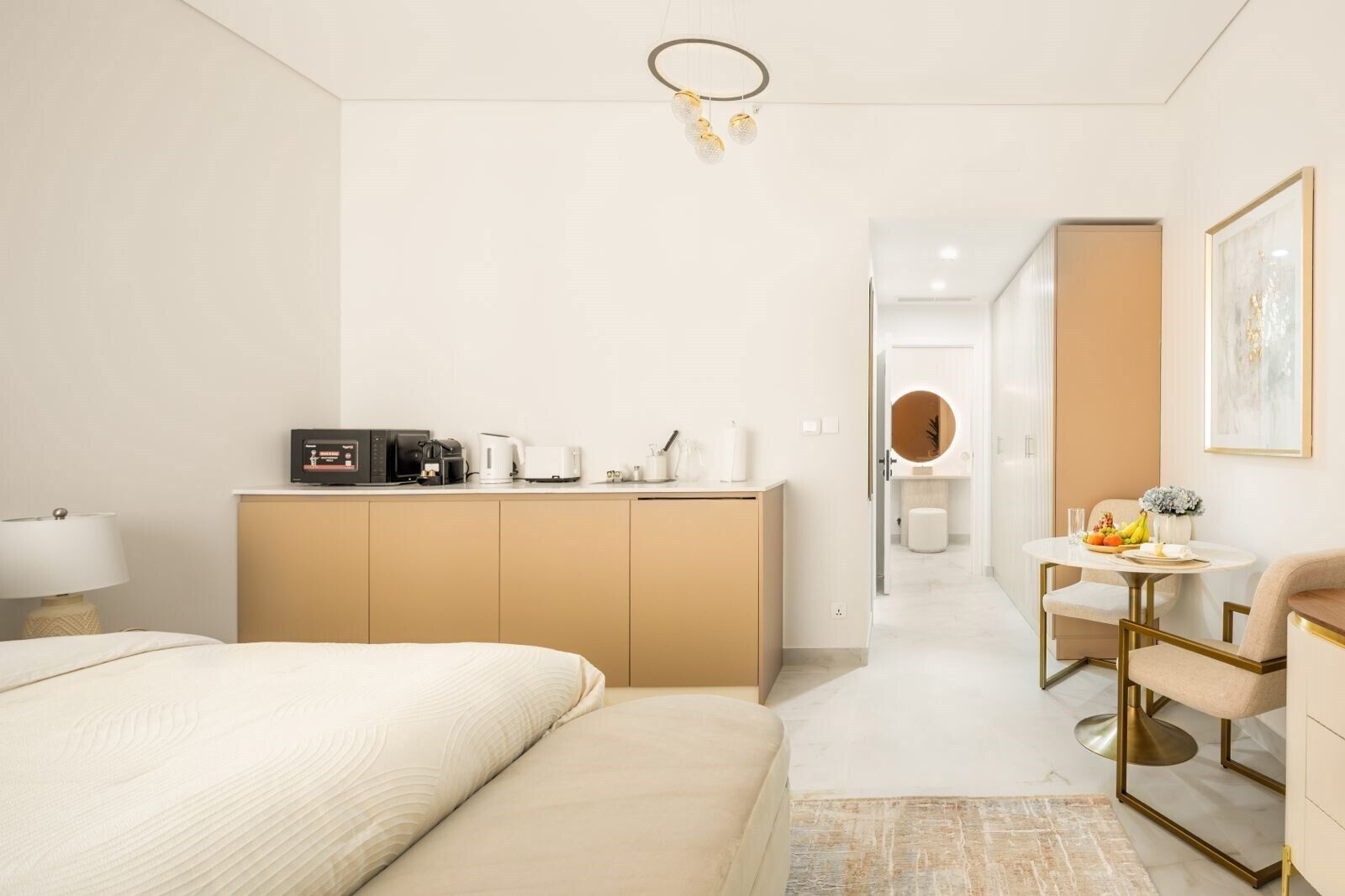 A modern hotel room with a bed, a desk with a coffee maker, a mirror, and a dining area with a table and chairs.