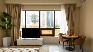 A modern hotel room with a large window, a flat-screen TV, a dining area with a table and chairs, and a potted plant.