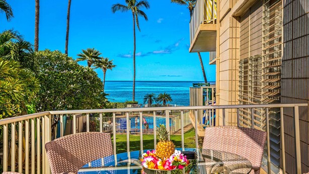 A balcony with wicker chairs and a glass table, overlooking a pool and palm trees, with a view of the ocean. - Oceanfront Kihei Condo With On-site Beach Access! (Kihei)