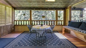 Une terrasse couverte avec un banc en bois, une table ronde recouverte d’une nappe à motifs et des jardinières suspendues.
