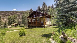 Une cabane en bois dotée d’un porche, entourée d’une for êt et d’un champ herbeux.