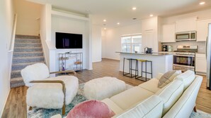 A modern living room with a sofa, ottomans, and a TV. A kitchen with white cabinets and appliances is visible in the background. - 12 Mi to Dtwn Austin: Quiet Home w/ Patio & Yard (Del Valle)