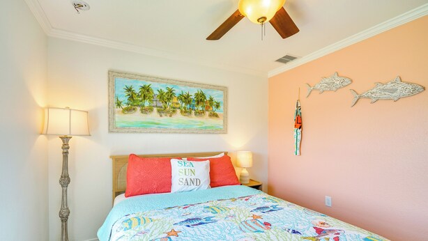 A bedroom with a bed, a nightstand, a floor lamp, a ceiling fan, a painting of a beach scene, and two wall-mounted fish decorations. - Pool w/ Waterfall: Colorful Cape Coral Gem! (Cape Coral)