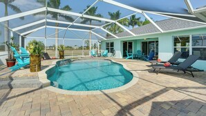 A covered swimming pool with lounge chairs, a small building with large windows, and palm trees in the background. - Pool w/ Waterfall: Colorful Cape Coral Gem! (Cape Coral)