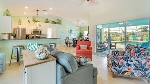 A spacious living area with a kitchen, a red armchair, a floral sofa, and a large glass door leading to an outdoor area. - Pool w/ Waterfall: Colorful Cape Coral Gem! (Cape Coral)