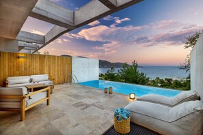 Un área de piscina moderna al aire libre con vista al mar, mobiliario de madera y cielo despejado.