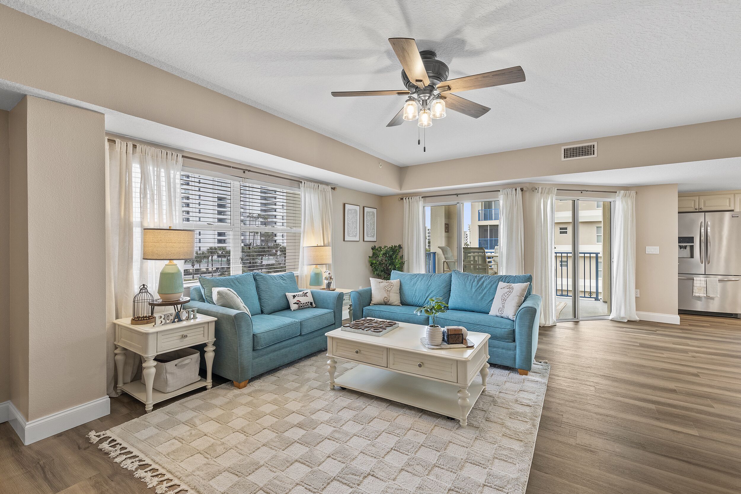 A spacious living room with a ceiling fan, a blue sofa set, a coffee table, and a view to the outside.