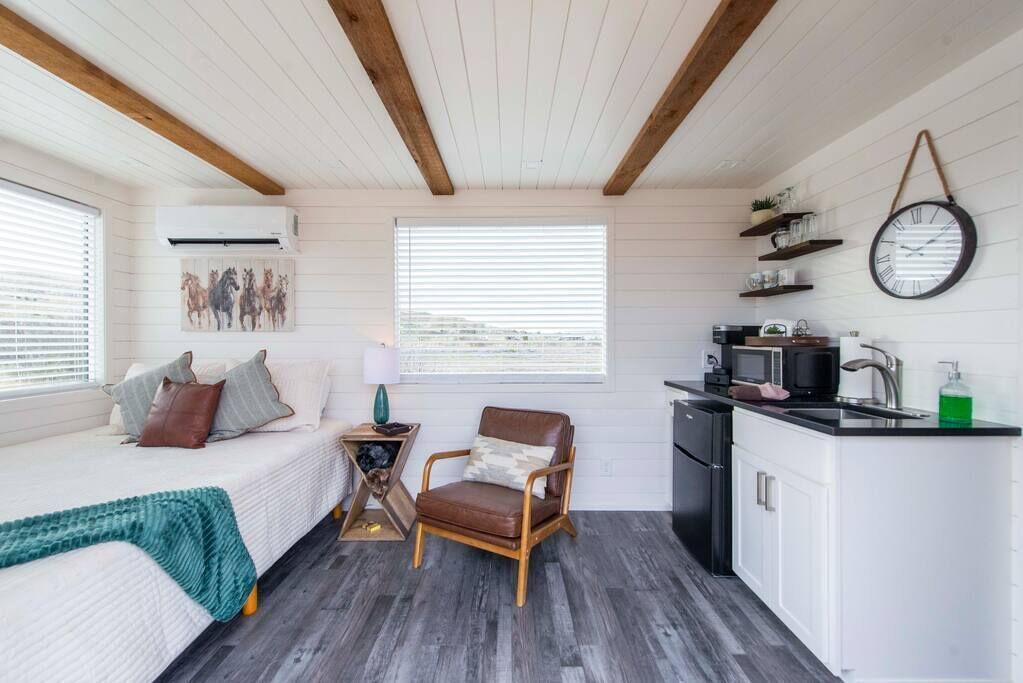 Une pièce de taille modeste comprenant un lit, une chaise, une petite table, un coin cuisine et une fenêtre avec des stores.