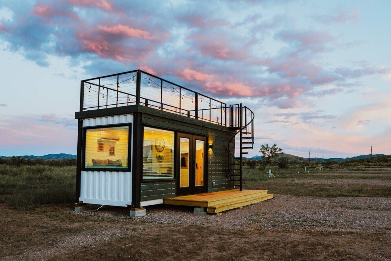 Una cabaña moderna de dos pisos con cerramiento de vidrio, una terraza de madera y una escalera en espiral.