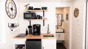A compact kitchen with a microwave, coffee maker, and a sink. There are shelves above the counter holding dishes and a telescope.