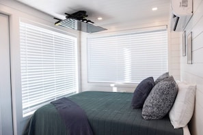 Une chambre à coucher avec un lit, deux fenêtres équipées de stores, un ventilateur de plafond et un appareil de climatisation.