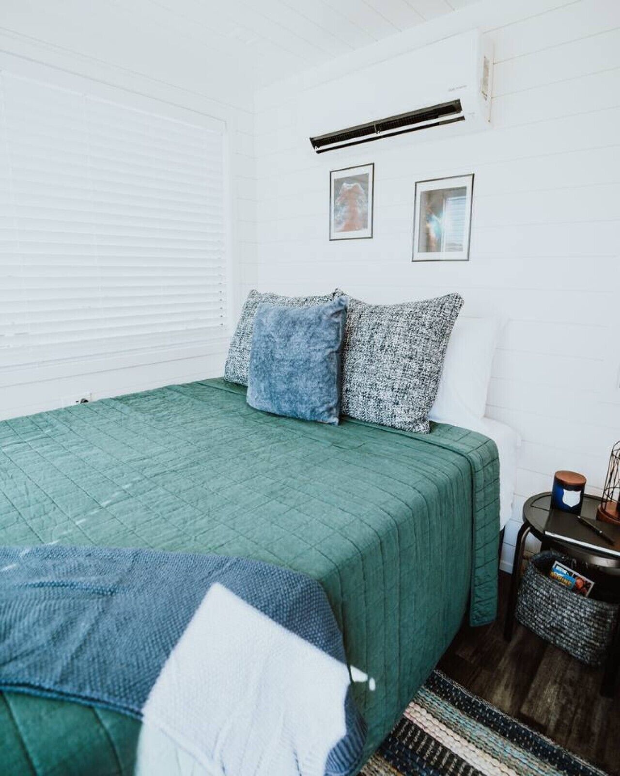 Una habitación con una colcha verde, dos almohadas, una mesita con una vela y un cubo de basura.