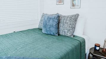 Una habitación con una colcha verde, dos almohadas, una mesita con una vela y un cubo de basura.