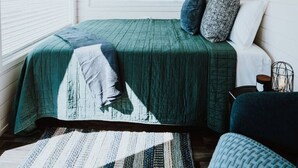 A room with a bed, a chair, a small table, and a window with blinds.
