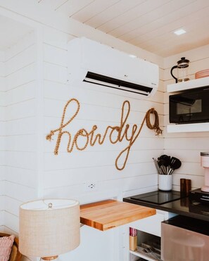 A kitchen with white walls, a hanging rope letter art, an air conditioner, a microwave, and a coffee maker. - New! Cowgirl Shipping Container Home (Alpine)